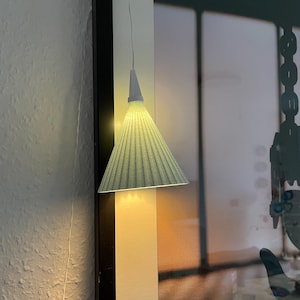 Puede incluir: Una pequeña lámpara colgante en forma de cono con una pantalla plisada de color verde claro cuelga de un fino cable blanco. La lámpara está colocada cerca de una obra de arte enmarcada, proyectando un brillo cálido sobre la pared. El diseño de la lámpara es minimalista y moderno.