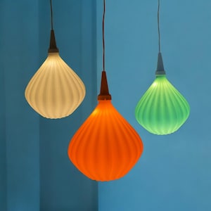 May include: Three pendant lights with a ribbed, teardrop-shaped design. One is white, one is orange, and one is green. Each light hangs from a brown cord and has a dark brown and gray top. The background is a solid blue.