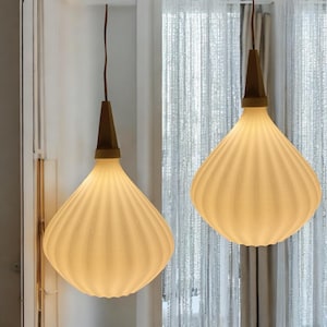 Puede incluir: Dos lámparas colgantes con un brillo cálido. Cada lámpara tiene una pantalla blanca, acanalada, en forma de lágrima y una parte superior de madera marrón. Las luces cuelgan de cables marrones, creando un ambiente suave y acogedor. El fondo incluye una cortina blanca.