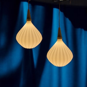 Puede incluir: Dos lámparas colgantes con pantallas en forma de lágrima, acanaladas y de color crema. Las lámparas tienen una parte superior de madera y cuelgan de un cable fino. El fondo es azul oscuro.