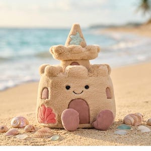 May include: A plush sandcastle toy in shades of tan and beige, with a smiling face and a star-shaped detail on top. The toy has a door and windows, and is surrounded by seashells and sea glass on a sandy beach.