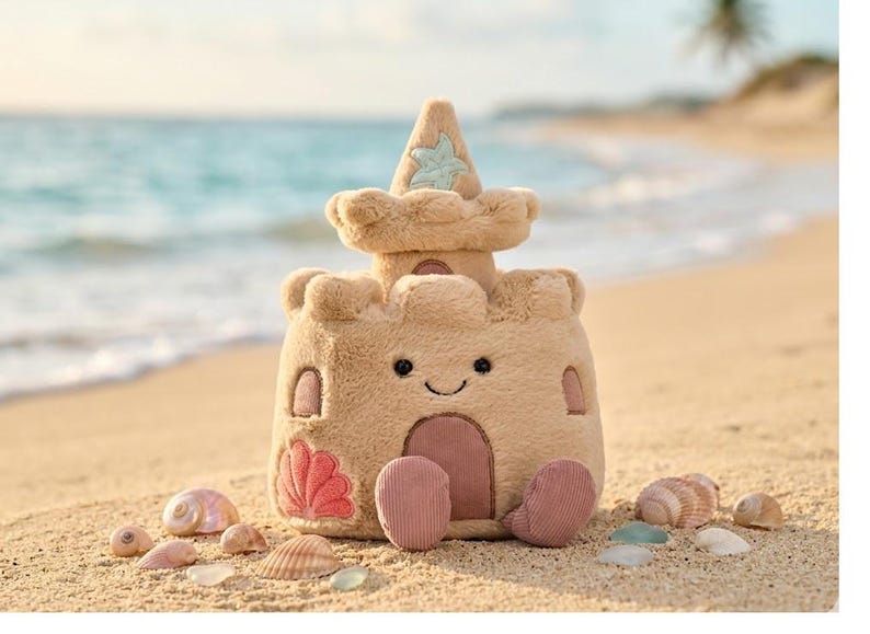 May include: A plush sandcastle toy in shades of tan and beige, with a smiling face and a star-shaped detail on top. The toy has a door and windows, and is surrounded by seashells and sea glass on a sandy beach.