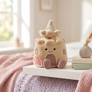 May include: A plush sandcastle toy in shades of tan, pink, and light blue. The toy has a smiling face, a door, and a star on top. It is sitting on a white tray with a vase and books.