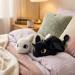 May include: Two plush dragon toys, one white and one black, rest on a bed with pillows and a blanket. The white dragon has blue eyes, while the black dragon has yellow eyes. The bed has a pink comforter and a light brown blanket.