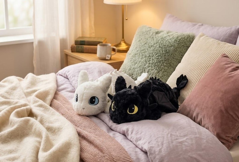 May include: Two plush dragon toys, one white and one black, rest on a bed with pillows and a blanket. The white dragon has blue eyes, while the black dragon has yellow eyes. The bed has a pink comforter and a light brown blanket.