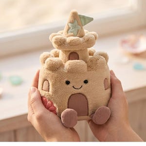 May include: A plush sandcastle toy in shades of tan and beige, featuring a smiling face, arched windows, and a decorative top with a flag. The toy has a soft, textured appearance and is designed to resemble a real sandcastle.