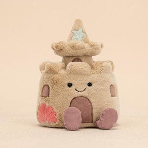 May include: A plush sandcastle toy in shades of tan and beige, featuring a smiling face, a pink seashell, and two pink corduroy feet. The sandcastle has a small tower with a light blue starfish design.