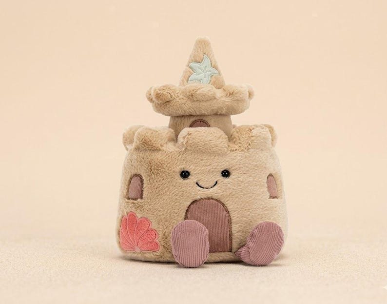May include: A plush sandcastle toy in shades of tan and beige, featuring a smiling face, a pink seashell, and two pink corduroy feet. The sandcastle has a small tower with a light blue starfish design.