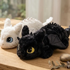 May include: Two plush dragon toys, one white and one black, rest on a wooden surface. The white dragon has blue eyes, while the black dragon has yellow eyes. Both have soft, textured fur and dragon-like features.
