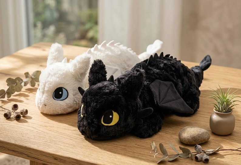 May include: Two plush dragon toys, one white and one black, rest on a wooden surface. The white dragon has blue eyes, while the black dragon has yellow eyes. Both have soft, textured fur and dragon-like features.