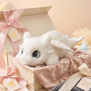 May include: A white plush dragon toy with blue eyes rests in an open gift box lined with pink velvet. The box is tied with pink ribbons and a tag. Decorative elements include a compass and small ornaments.