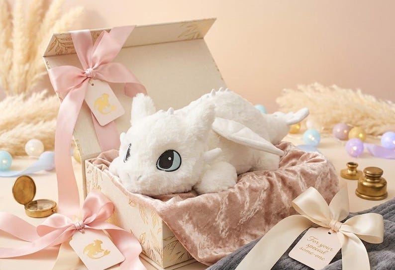 May include: A white plush dragon toy with blue eyes rests in an open gift box lined with pink velvet. The box is tied with pink ribbons and a tag. Decorative elements include a compass and small ornaments.