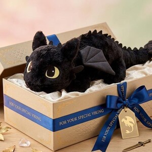 May include: A black plush dragon toy with large yellow eyes, wings, and a spiky spine, presented in a decorative gift box. The box is beige with a blue ribbon that reads "FOR YOUR SPECIAL VIKING". A gold tag with a dragon silhouette is attached.