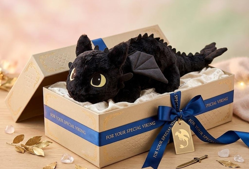May include: A black plush dragon toy with large yellow eyes, wings, and a spiky spine, presented in a decorative gift box. The box is beige with a blue ribbon that reads "FOR YOUR SPECIAL VIKING". A gold tag with a dragon silhouette is attached.