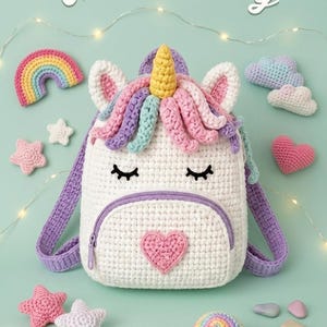 Pode incluir: Uma mochila de unicórnio de crochê branca com zíper e alças roxas, adornada com um chifre amarelo, crina rosa e roxa e olhos fechados. A imagem inclui estrelas de crochê, um arco-íris, nuvens e um coração.