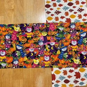 May include: A rectangular fabric runner featuring a vibrant Halloween print with cartoon cats dressed in witch hats and costumes amidst orange pumpkins on a black background, contrasted by white end panels patterned with colorful autumn leaves.