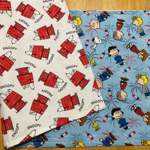 May include: A double-sided fabric runner featuring two distinct patterns. One side is white with a repeating print of Snoopy lying on a red doghouse and the text Snoopy. The other side is light blue with a pattern of Peanuts characters and fireworks.