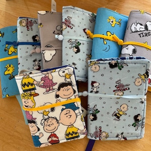May include: A collection of fabric-covered notebooks featuring Peanuts characters. The notebooks are in various colors, including blue, gray, and white, and are secured with elastic bands. The designs include Snoopy, Charlie Brown, and other characters.