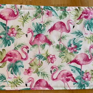 May include: A white fabric table runner with a pink flamingo and tropical leaf pattern. The runner has a stitched edge.