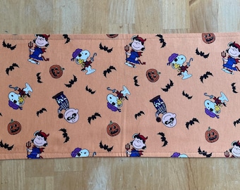 May include: Rectangular orange placemat with a repeating Halloween-themed pattern. The design features cartoon characters, bats, and jack-o'-lanterns. The placemat has a stitched border.