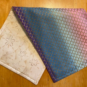 May include: A reversible table runner featuring two distinct patterns. One side showcases a cream-colored fabric with a repeating sand dollar design in a muted gold. The other side displays a gradient of blue, purple, and pink with a gold scale pattern.