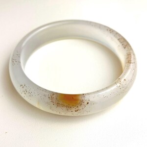 May include: A translucent, smooth, circular bangle bracelet made of light gray stone. The material features natural inclusions, including scattered dark brown speckles and a single, prominent, circular amber-colored patch surrounded by a dark ring.