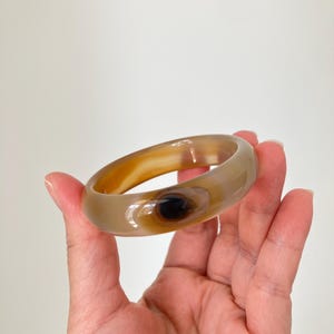 Puede incluir: Un brazalete rígido liso y pulido, elaborado en piedra ágata translúcida de color marrón claro. La banda circular presenta una distintiva inclusión en forma de ojo de color marrón oscuro en un lado, rodeada por anillos concéntricos en tonos beige.