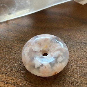 May include: A circular, donut-shaped pendant made of translucent, polished stone. The material features a pale, milky white base with intricate, flower-like inclusions in shades of soft pink and peach, centered around a small, smooth, circular hole.