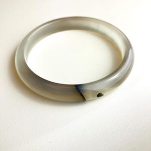 以下が含まれることがあります： 淡いグレーとベージュの石で作られた、滑らかで半透明のバングルブレスレット。円形のバンドには、自然な暗いインクルージョンと縞模様がかすかに見られ、磨かれた表面全体に柔らかな大理石のような外観を作り出しています。