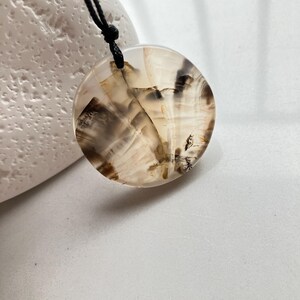May include: A circular pendant featuring a translucent, polished stone with natural inclusions of dark brown and black dendritic patterns. The pendant is suspended from a thin, black cord that passes through a small hole at the top of the stone.