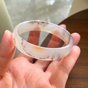 以下が含まれることがあります： 滑らかに磨かれた石で作られた半透明の円形バングルブレスレット。素材は乳白色のベースに、かすかな曇った灰色のインクルージョンがあり、外縁に沿って温かみのある琥珀色の小さな斑点がはっきりと見られます。