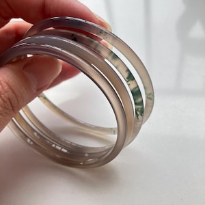 Puede incluir: Tres brazaletes circulares y lisos fabricados en piedra translúcida de color gris claro. Los brazaletes presentan sutiles inclusiones verdes similares al musgo dispersas por el material, creando un patrón natural y variado en cada pieza.