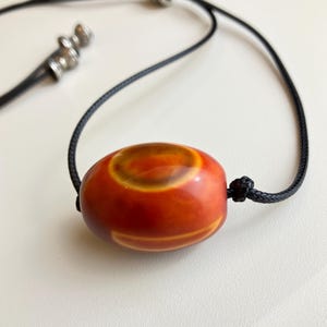 Könnte beinhalten: Eine glatte, oval geformte Perle mit polierter Oberfläche in gebrannten Orange- und Dunkelrottönen, die ein markantes gelbes kreisförmiges Muster aufweist. Die Perle ist auf einer dünnen schwarzen Kordel mit kleinen, silberfarbenen Endkappen.