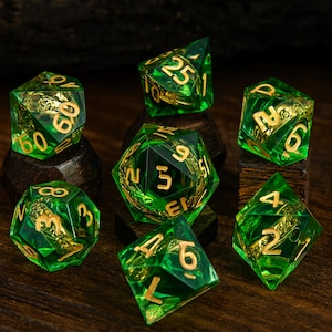 May include: A collection of seven green and gold polyhedral dice. The dice are transparent with gold numbering and internal designs. They are arranged on small, dark wooden blocks against a dark wood background. The dice are used for tabletop games.