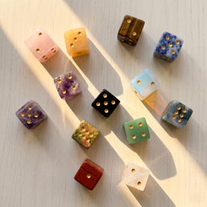 Puede incluir: Una colección de doce dados de piedras preciosas en varios colores y patrones. Los dados incluyen cuarzo rosa, ámbar, ojo de tigre, sodalita, amatista, ónix negro, ópalo y otros. Cada dado tiene puntos dorados.
