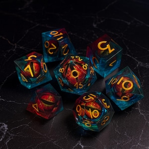 May include: Set of seven polyhedral dice with a fantasy theme. Each die is transparent blue with a red and yellow eye design inside. The numbers are gold. Perfect for tabletop role-playing games.