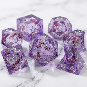 May include: Set of seven polyhedral dice in various shapes, each with a transparent purple resin body. The dice feature silver numbering and are adorned with pink floral inclusions. Ideal for tabletop gaming.