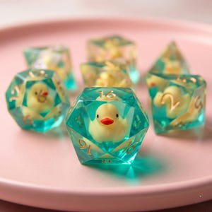 May include: A set of polyhedral dice with a whimsical design. Each die is a clear, teal-colored resin, encasing a small, yellow duck with an orange beak. The dice are displayed on a pink plate.