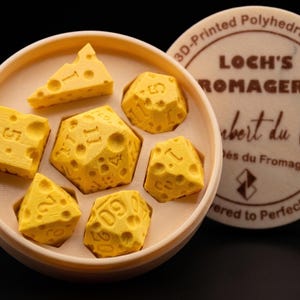Op de afbeelding: Een set gele, kaasvormige polyhedrale dobbelstenen in een ronde container. De dobbelstenen zijn 3D-geprint en hebben ingegraveerde cijfers. De container is beige en het deksel leest "LOCH'S ROMAGERIE" en "3D-Printed Polyhedral Diceets."