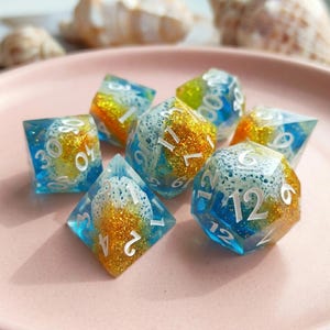 Könnte beinhalten: Set aus sieben polyedrischen Würfeln mit einem Meeres-Design. Die Würfel sind transparent mit blauen, goldenen und weißen Akzenten und weißen Zahlen. Für Tischspiele.
