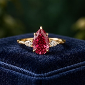 May include: A gold ring featuring a large pear-cut red gemstone as the center stone, flanked by two smaller pear-cut clear white stones on each side, all set in a polished yellow gold band with a prong setting design.