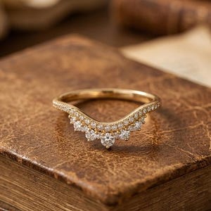 Pode incluir: Um anel de ouro em forma de chevron com uma fila de pequenos diamantes em pavé ao longo da banda e uma série de diamantes redondos maiores dispostos em forma de V ao longo da borda inferior, criando um visual em camadas e brilhante.