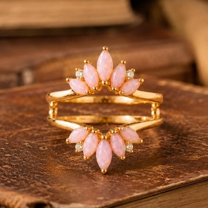 May include: A gold ring featuring a double-band design with two arched sections. Each section is adorned with a row of marquise-cut pink stones and small, clear round accent stones arranged in a fan-like pattern along the top and bottom.