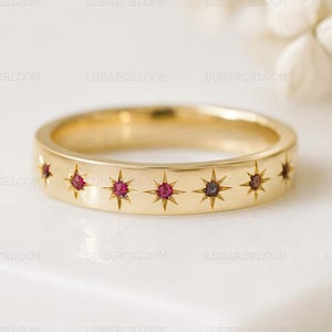 May include: A gold-colored ring with a series of star-shaped cutouts, each containing a small, round, red gemstone. The ring is set against a white background, showcasing its intricate design and craftsmanship. The ring is a piece of jewelry.
