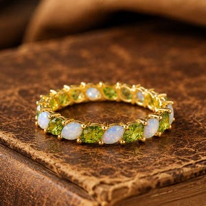 Puede incluir: Un anillo de banda de oro con un patrón alterno de ópalos blancos ovalados y piedras preciosas de peridoto verde de corte redondo en un estilo de eternidad continuo. El engaste es de oro amarillo pulido con una secuencia repetida.