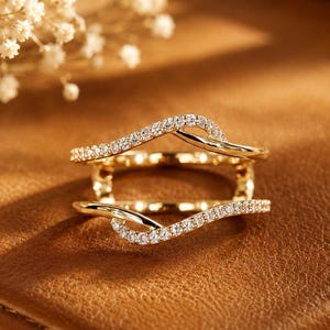 May include: A gold-colored ring with a unique design, featuring two curved bands encrusted with small, sparkling stones. The ring sits on a brown leather surface, with a sprig of white flowers in the background.