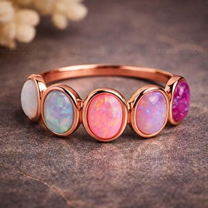 May include: A rose gold ring featuring five oval-shaped opals in shades of white, blue, pink, and purple. The gemstones are set in a row, creating a colorful and elegant design. The ring is a piece of jewelry.