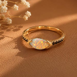May include: A gold ring featuring an oval opal gemstone, displaying iridescent colours. The ring is flanked by small black stones. The ring is set against a brown backdrop, with white flowers in the background.