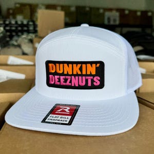 May include: White flat bill snapback hat with a mesh back. The front features a patch with the words "DUNKIN' DEEZNUTS" in orange and pink. A black and red tag is attached to the brim.