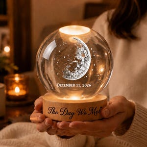 May include: A clear glass sphere lamp featuring an internal laser-etched crescent moon and stars, with the text "DECEMBER 11, 2026" printed on the glass and "The Day We Met" engraved on the circular light-colored wooden base.